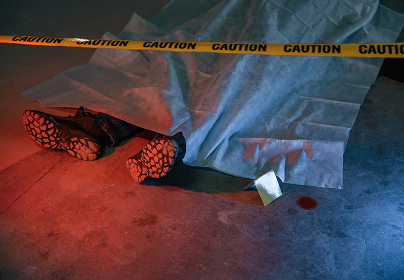Covered body on the ground with black boots, surrounded by caution tape and a small pool of blood.