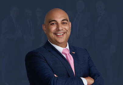 Smiling man in a navy suit and pink tie, arms crossed, with a dark background featuring blurred figures.