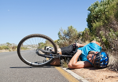 Cyclist in a blue helmet and tank top lies on the ground beside a bike on a sunny road with shrubs.