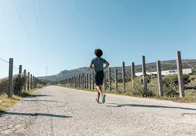 A person with curly hair jogs along a gravel path lined with wooden posts and mountains in the background.