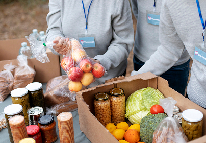 Volunteers in gray sweatshirts pack fresh produce, jars, and canned goods into cardboard boxes at an outdoor food drive.