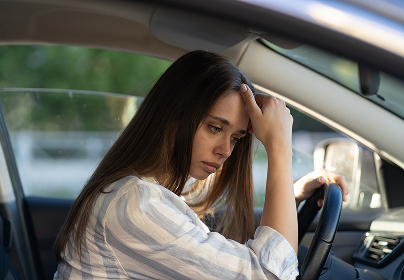 Woman with long brown hair looks distressed while sitting in a car, resting her forehead on her hand.