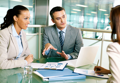Three professionals in business attire discuss documents and a laptop in a modern office setting.