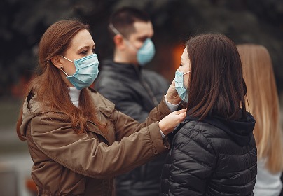 Two women adjust each other's blue masks outdoors, with two men in the background also wearing masks.