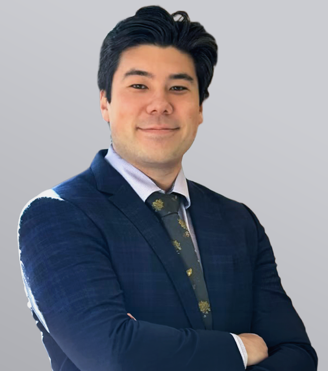 Man in a navy suit with a patterned tie, smiling confidently against a gray background. Arms crossed.